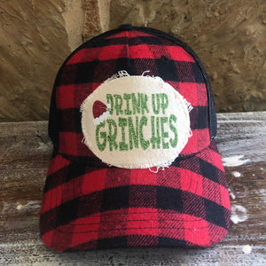 Red and black checkered cap with 'Drink Up Grinches' patch on a wooden surface.