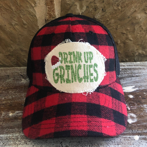 Red and black checkered cap with 'Drink Up Grinches' patch on a wooden surface.