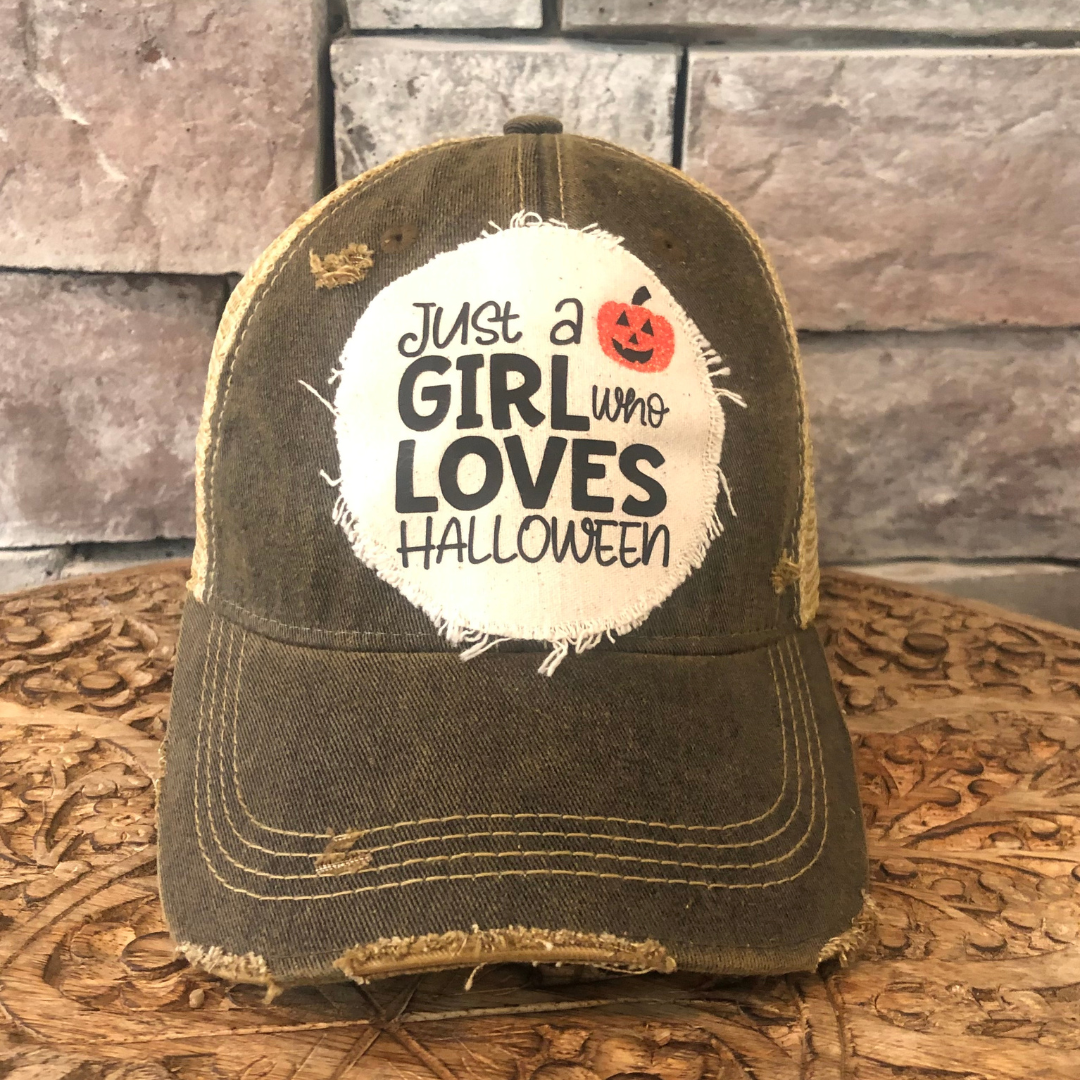 Distressed brown cap with 'Just a Girl Who Loves Halloween' text and pumpkin graphic on a wooden surface.