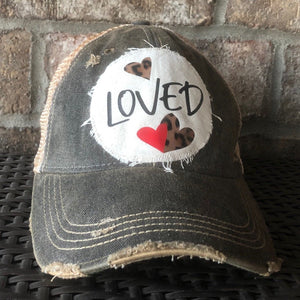 A distressed, vintage feel baseball hat in black with an oversized white 'LOVED' logo decorated with a small leopard print and red hearts.