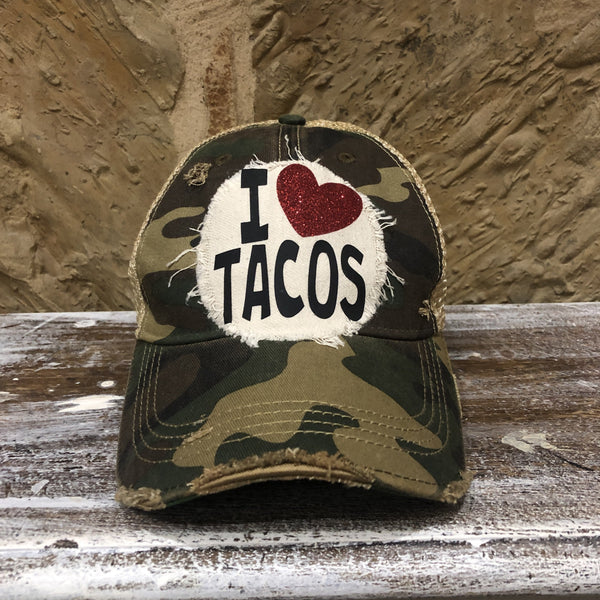 A camouflage baseball hat with an oversized 'I ❤️ TACOS' logo in white and black, featuring a glitter heart.